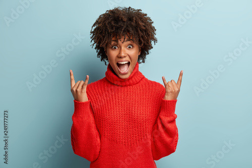 Charismatic lady raises hands with rock n roll gesture, feels glad while dances at party, listens favourite music, keeps jaw dropped, wears casual winter outfit, isolated over blue background