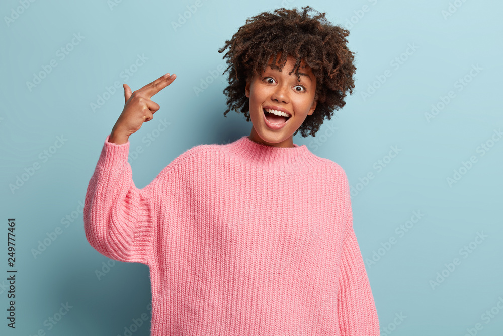 Positive ethnic woman with curly hair, dressed in pink jumper, makes suicide gesture, smiles and has fun, models over light blue background being crazy, pretends killing herself and shooting in temple Positive ethnic woman with curly hair, dressed in pink jumper, makes suicide gesture, smiles and has fun, models over light blue background being crazy, pretends killing herself and shooting in temple