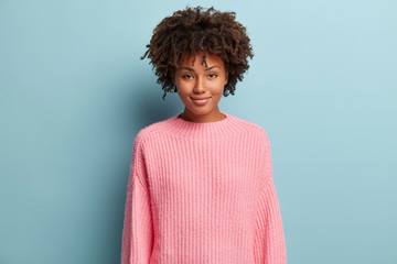 Portrait of attractive black woman with curly hairstyle, dressed in pink sweater, looks with enjoyment at camera, has happy expression, isolated over blue background. Ethnicity and beauty concept