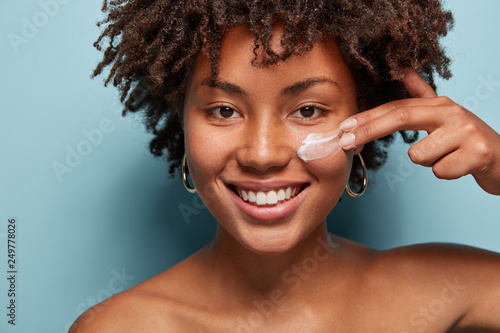 Close up shot of positive dark skinned woman with curly hair, appiles face cream on cheek, poses nude against blue background, cares of beauty, uses cosmetic product Fototapet