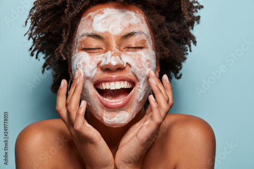 Portrait of beautiful woman with white foam mask on face, touches cheeks, enjoys softness of skin after beauty treatments, has curly hair, shows bare shoulders, isolated over blue background