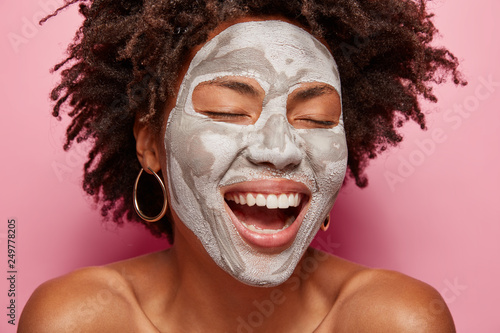 Face care concept. Smiling positive woman with white clay mask, has pore cleaning procedure, laughs happily, shows white even teeth, has crisp hair, poses bare shoulders over pink background
