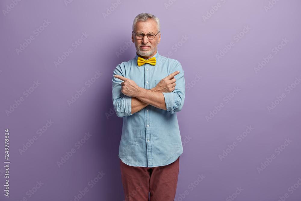 Indoor shot of handsome hesitant grandfather crosses arms over chest, points with fore fingers at different empty corners, makes decision, wears fashionable clothes, isolated over purple background Indoor shot of handsome hesitant grandfather crosses arms over chest, points with fore fingers at different empty corners, makes decision, wears fashionable clothes, isolated over purple background