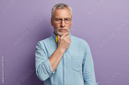 Headshot of attractive wrinkled man with grey hair holds chin, contemplates about life, dressed in stylish shirt, wears spectacles, poses over purple background. People, age, experience concept