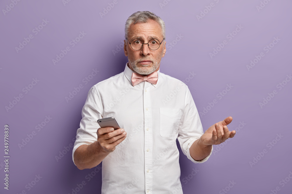 Image of handsome mature man with grey short hair, wears bowtie and shirt, gestures with hand, holds mobile phone, puzzled to recieve text message from stranger, isolated over purple background Image of handsome mature man with grey short hair, wears bowtie and shirt, gestures with hand, holds mobile phone, puzzled to recieve text message from stranger, isolated over purple background