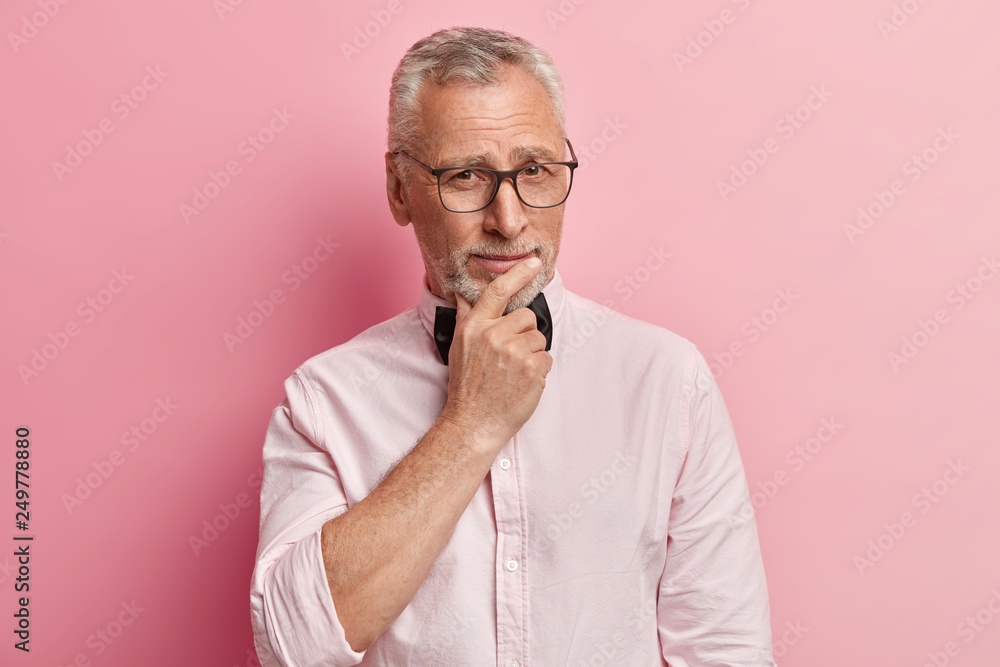 Headshot of serious mature Caucasian man holds chin, looks with eyes full of wisdom, dressed in elegant clothes, isolated over pink studio background, wants to share his experience with grandson. Headshot of serious mature Caucasian man holds chin, looks with eyes full of wisdom, dressed in elegant clothes, isolated over pink studio background, wants to share his experience with grandson.