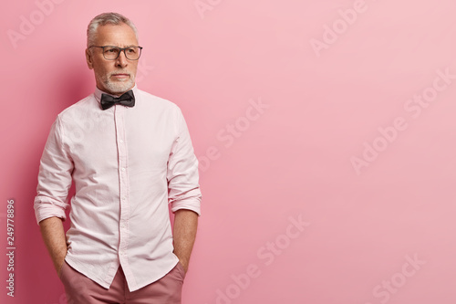 Half length shot of attractive senior man with grey hair and beard, keeps hands in pockets of trousers, wears elegant shirt and bowtie, being deep in thoughts, isolated over pink background. © wayhome.studio 