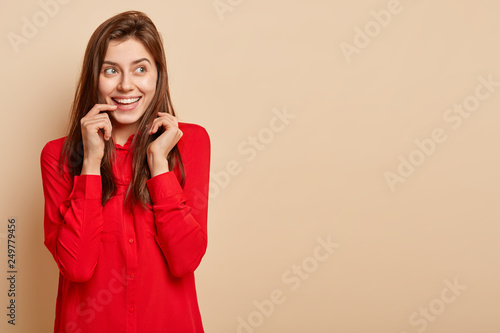 Image of pretty lady smiles gently, keeps finger near lips, focused aside, dressed in red shirt, models against beige background with copy space, has dreamy expression. Emotions, beauty concept