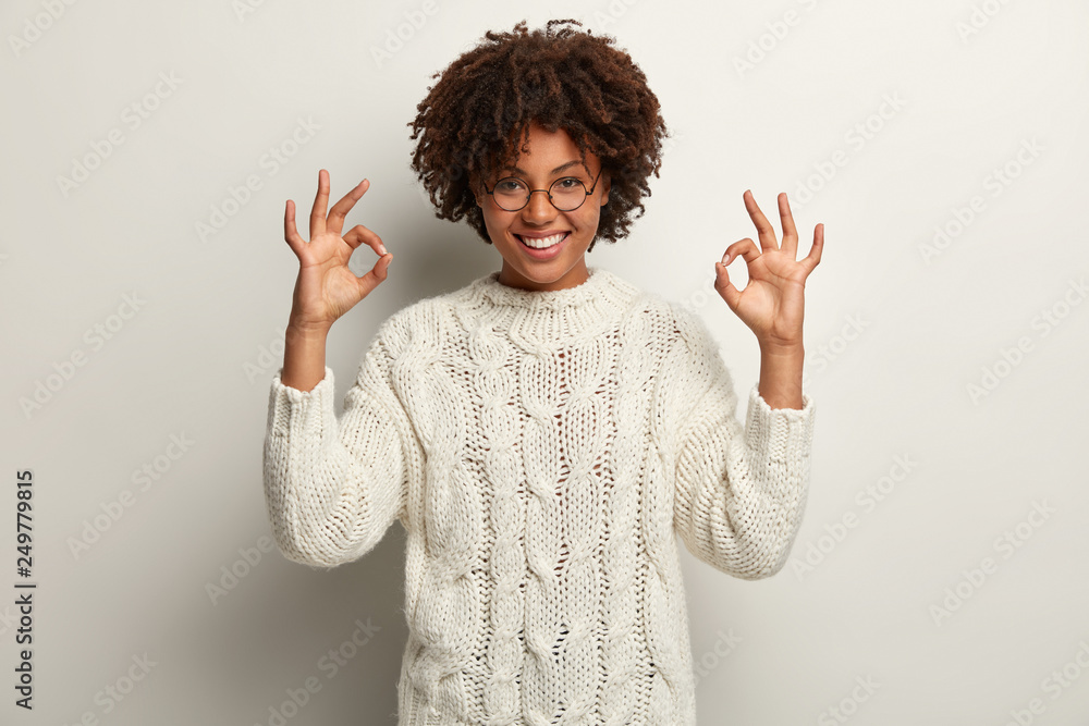 Approval and agreement concept. Picture of happy black ethnic woman makes okay gesture with both hands, says everything is super or perfect, dressed in white jumper, proceeds according to plan Approval and agreement concept. Picture of happy black ethnic woman makes okay gesture with both hands, says everything is super or perfect, dressed in white jumper, proceeds according to plan
