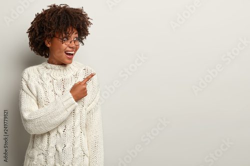 Positive pleased young woman with crisp hair, points with fore finger aside, wears spectacles and sweater, shows something over blank space, being in good mood. Ethnicity and promotion concept