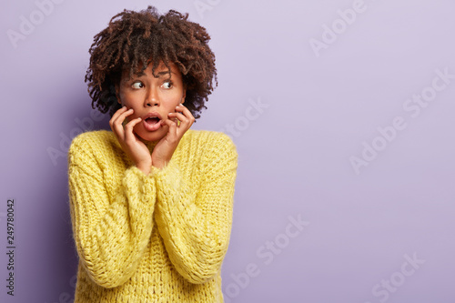 Oh no  I am afraid of this. Puzzled black woman has bushy hairstyle  keeps mouth opened from amazement  looks aside with fright  dressed in yellow knitted sweater  blank space over purple background