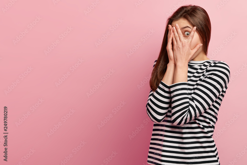 Photo of surprised woman has dark long hair, peeps through fingers with amazement, feels shocked and shy, hides her face, wears striped jumper, isolated over rosy background with free space. Photo of surprised woman has dark long hair, peeps through fingers with amazement, feels shocked and shy, hides her face, wears striped jumper, isolated over rosy background with free space.