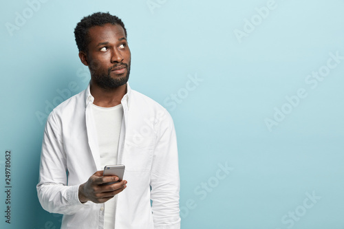 Pensive black man focused upwards, thinks what to write, has to give feedback, holds mobile phone, wears white shirt, isolated over blue studio wall with copy space, connected to wireless internet