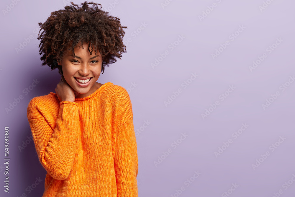 Upbeat tender young girlfriend with curly hairstyle, keeps hand on neck, wears bright outfit, smiles broadly, shows white teeth, models over purple background with empty space for your promotion Upbeat tender young girlfriend with curly hairstyle, keeps hand on neck, wears bright outfit, smiles broadly, shows white teeth, models over purple background with empty space for your promotion