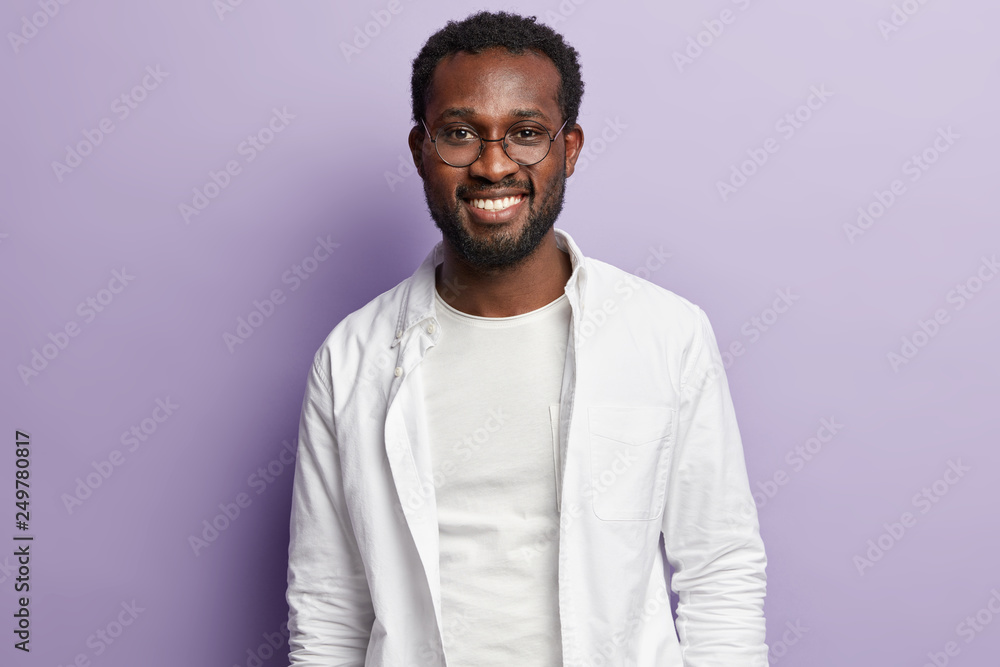 Photo of attractive black man with thick bristle, toothy smile, wears white stylish shirt, round spectalces, rejoices new task for accomplishment, isolated over purple background. Ethnicity concept Photo of attractive black man with thick bristle, toothy smile, wears white stylish shirt, round spectalces, rejoices new task for accomplishment, isolated over purple background. Ethnicity concept