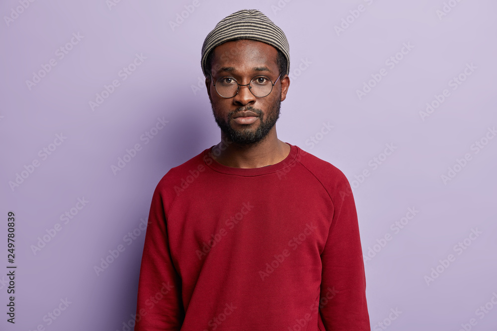 Hipster guy with black skin, looks seriously at camera, wears hat, spectacles and red jumper, listens advice of friend, has thick bristle, isolated over purple background. Ethnicity, youth concept Hipster guy with black skin, looks seriously at camera, wears hat, spectacles and red jumper, listens advice of friend, has thick bristle, isolated over purple background. Ethnicity, youth concept