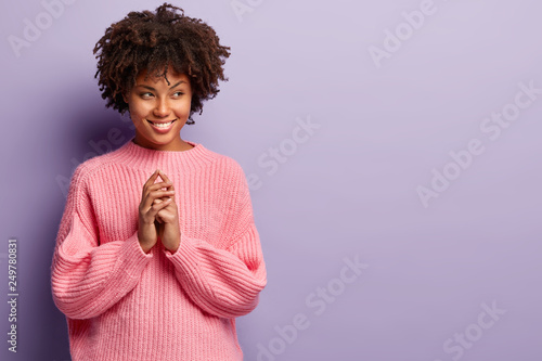 Positive dark skinned woman with toothy smile, dressed in oversized jumper, keeps hands together, has intention for something incredible, wears pink sweater, isolated over purple background.