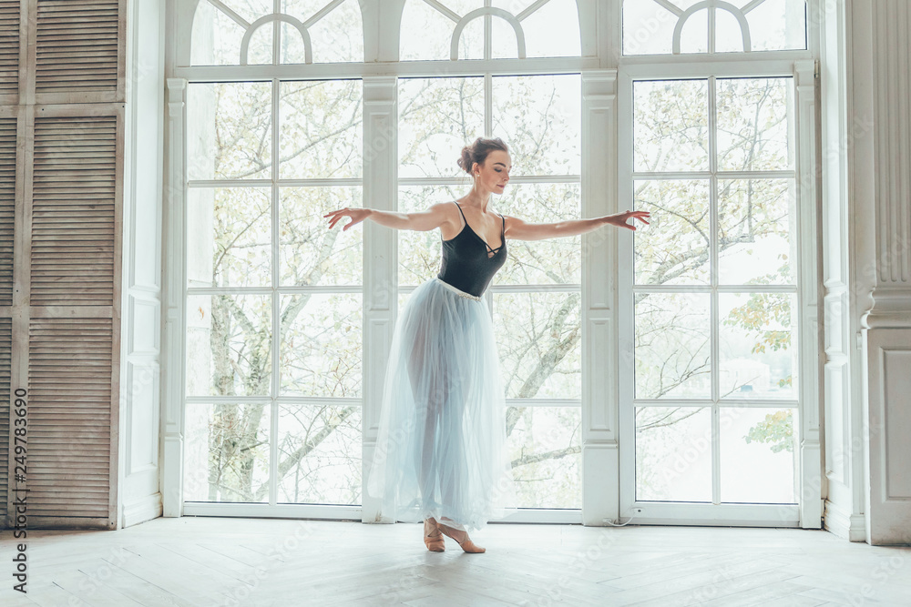 Young classical ballet dancer woman in dance class. Beautiful graceful ballerina practice ballet positions in blue tutu skirt near large window in white light hall Young classical ballet dancer woman in dance class. Beautiful graceful ballerina practice ballet positions in blue tutu skirt near large window in white light hall