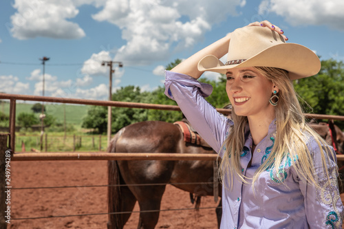 Beautiful blonde cowgirl with hat standing near the horse ranch background