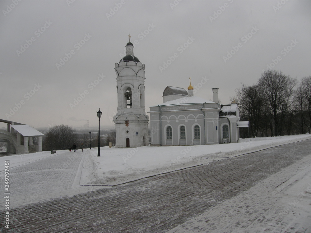 White Church with a bell tower on the background of low gray winter sky and snow-covered brick paths