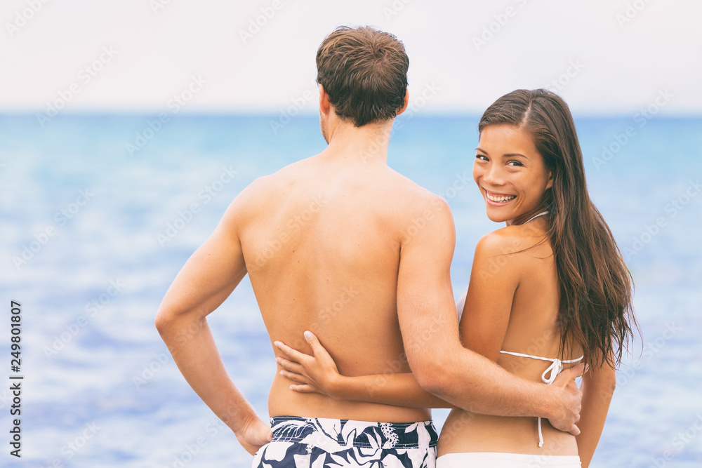 Happy couple hugging relaxing at the beach. Asian girlfriend looking back smiling at camera with boyfriend enjoying ocean view swimming in summer vacation destination. Happy couple hugging relaxing at the beach. Asian girlfriend looking back smiling at camera with boyfriend enjoying ocean view swimming in summer vacation destination.