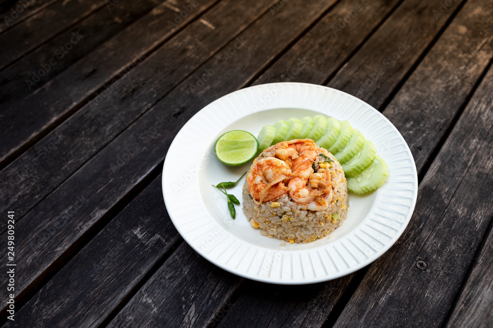 Thai style shrimps fried rice with fresh cucumber and lime on white ceramic plate on vintage wooden table background, healthy food