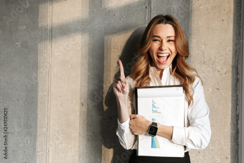 Beautiful young business woman dressed in formal clothes shirt indoors holding documents pointing.