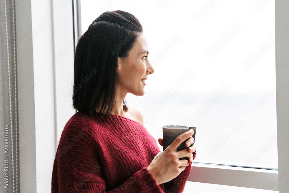 Pretty woman indoors in home near window drinking coffee. Pretty woman indoors in home near window drinking coffee.