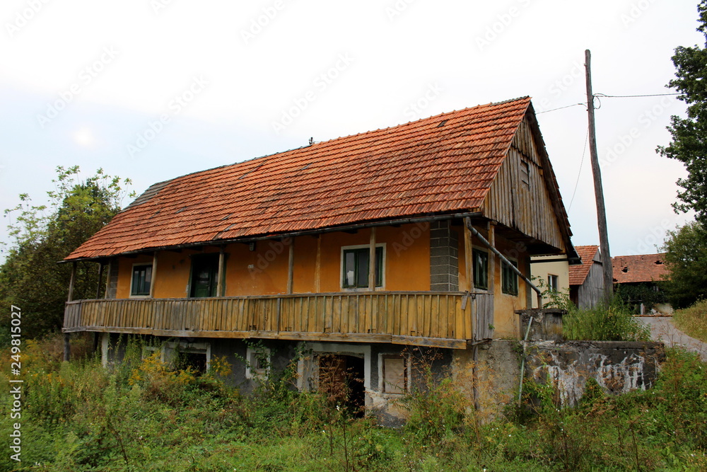 Abandoned stone and wooden family house with front porch surrounded with uncut grass and overgrown forest vegetation next to tall wooden utility pole and asphalt road Abandoned stone and wooden family house with front porch surrounded with uncut grass and overgrown forest vegetation next to tall wooden utility pole and asphalt road