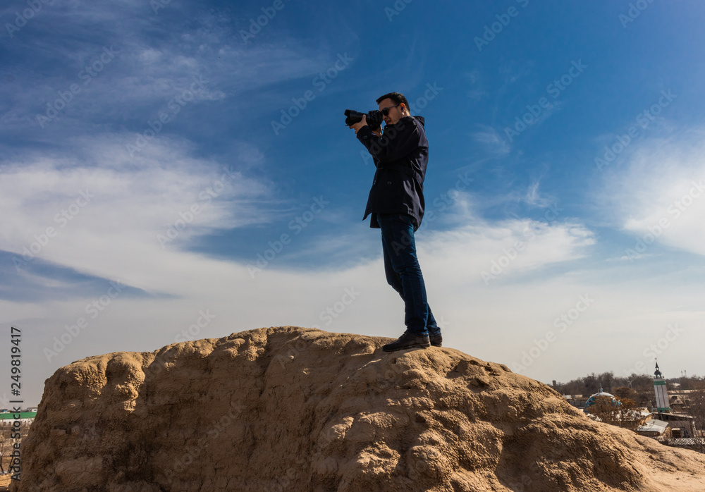 photographer in dark outfit and black sunglasses holding a camera steady in his hands and making a shot while standing on top of an ancient wall. Aerial view.
