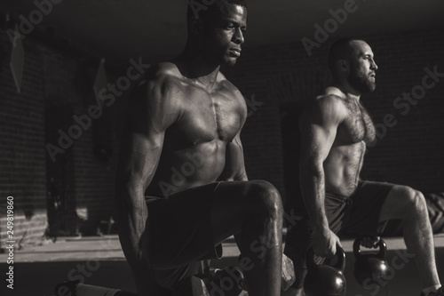 Black and white photo of two young caucasian and african male athletes with kettlebells making lunges with weight in indoor workout