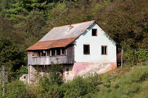 Dilapidated old family house with cracked facade and wooden balcony built on small hill surrounded with dense forest on warm summer day © hecos