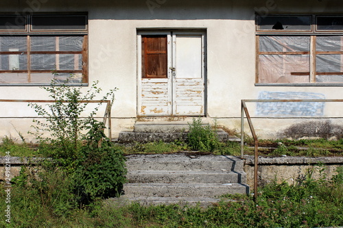 Front entrance to an abandoned house with destroyed windows and broken doors locked with strong padlock surrounded with overgrown grass and other plants covering concrete steps and rusted handrails on