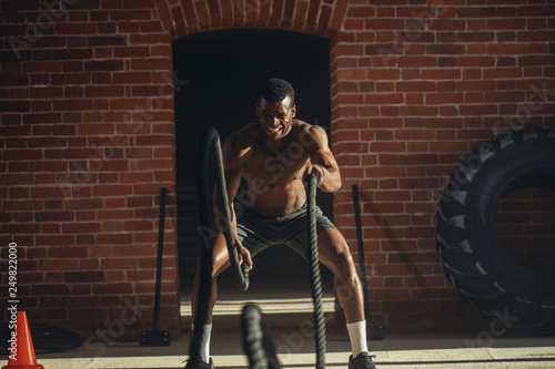 Muscular powerful determined african man training with rope in functional training in outdoor gym with brick walls, strengthen the muscles of the shoulders, which is important for boxers and swimmers