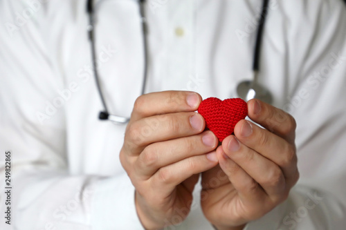 Cardiologist appointment, doctor with stethoscope holding red knitted heart in hands. Concept of cardiology, heart diseases, diagnosis, medical exam in clinic