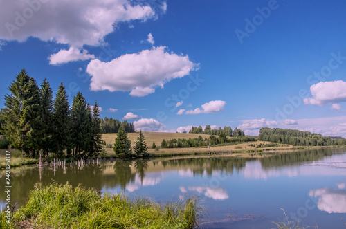 Summer view of the water surface of the river with overgrown picturesque banks and Cumulus clouds in the blue sky, toned.