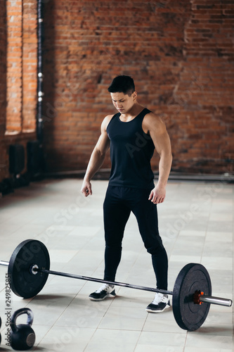 strong muscular guy is standing nera the barbell and looking at it. preparation for training, concentration before workout. full length photo.