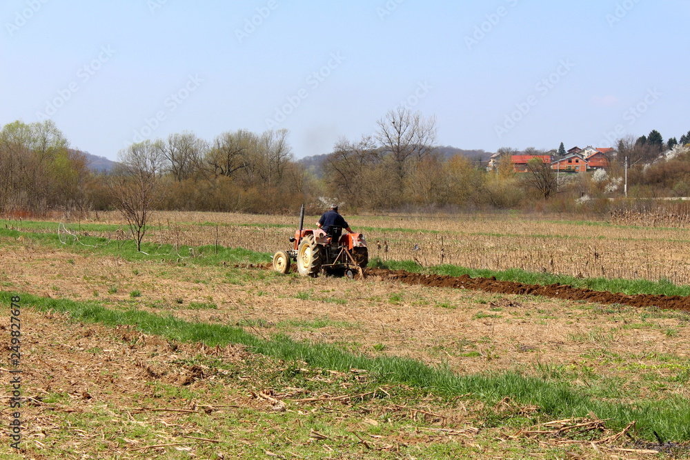 Obraz premium Open field with man on older tractor plowing in spring surrounded with uncut grass and cornfield with trees and family houses in background on warm sunny day