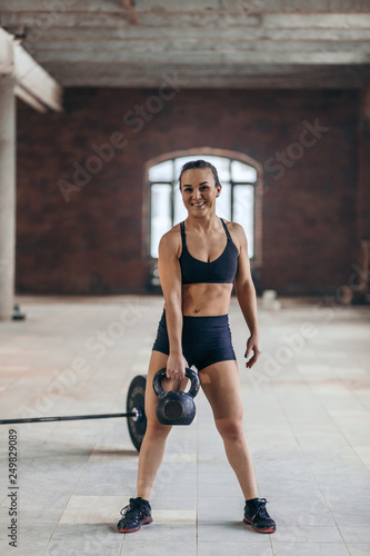 cheerful young sporty girl training with weight at gym. full length photo. happy sportswoman holding a kettlebell and looking at the camera