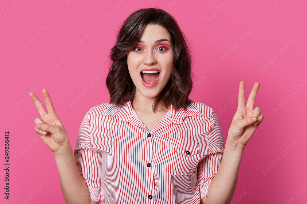 Optimistic fashionable woman with dark hair, smiles happily, makes peace gesture with both hands, dressed in stylish shirt, has fun indoor, isolated over pink background, has professional makeup Optimistic fashionable woman with dark hair, smiles happily, makes peace gesture with both hands, dressed in stylish shirt, has fun indoor, isolated over pink background, has professional makeup