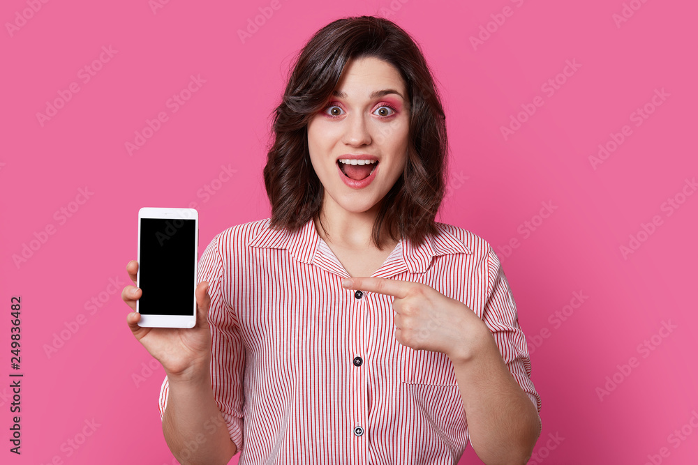 Portrait of independent, excited girl in pink, striped shirt with round buttons holding white mobile phone with black screen in fingers, and her second hand points to switched off smartphone. Portrait of independent, excited girl in pink, striped shirt with round buttons holding white mobile phone with black screen in fingers, and her second hand points to switched off smartphone.