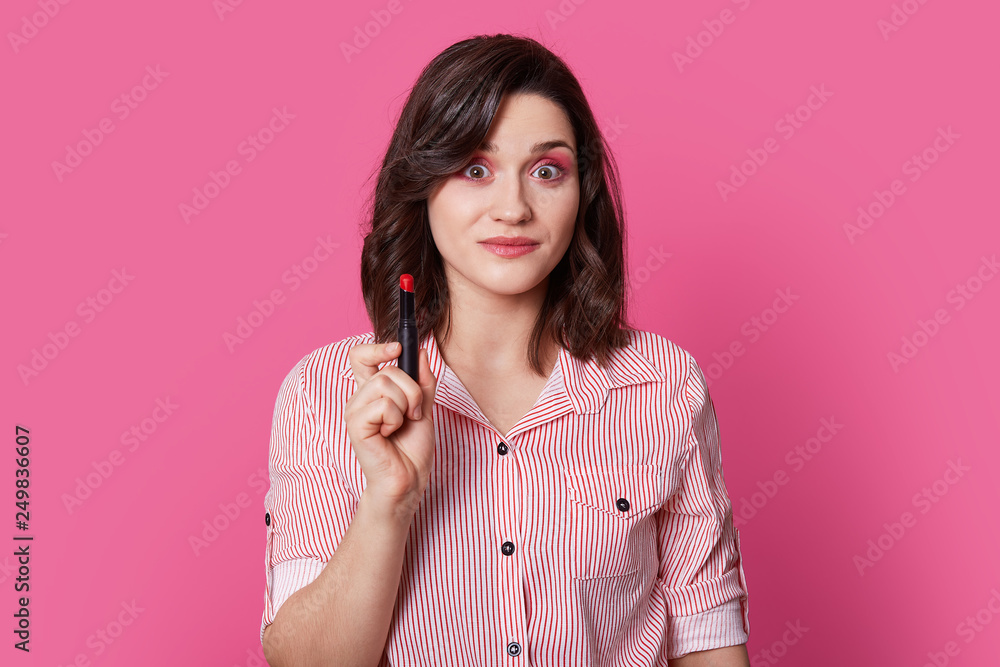 Photo of surprised brunette young woman with trendy hairstyle, has shocked expression, holds red lipstick, wears striped shirt, isolated over bright pink background. People and beauty concept Photo of surprised brunette young woman with trendy hairstyle, has shocked expression, holds red lipstick, wears striped shirt, isolated over bright pink background. People and beauty concept