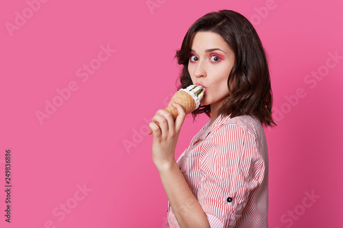 Sideways shot of attractive brunette lady licks ice cream, has professional make up, dressed in striped shirt, isolated over pink background with free space for your promotional content. Summer time