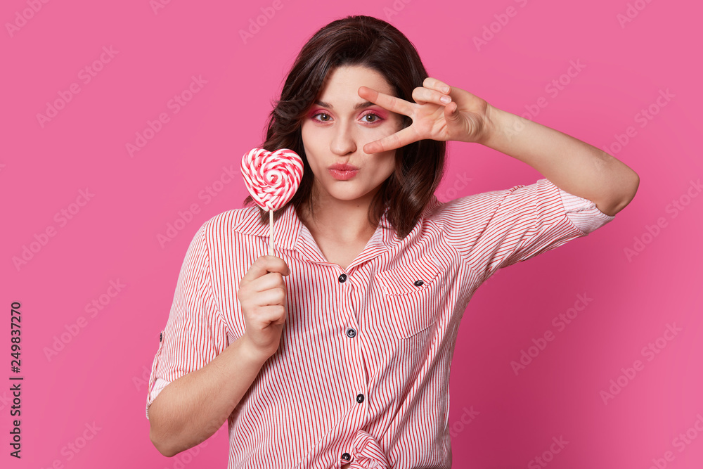 Horizontal view of attractive young woman has makeup, holds peace sign, holds candy, dressed in striped shirt, isolated over pink background, shows her beauty. Stylish coquetish girl with sweet Horizontal view of attractive young woman has makeup, holds peace sign, holds candy, dressed in striped shirt, isolated over pink background, shows her beauty. Stylish coquetish girl with sweet