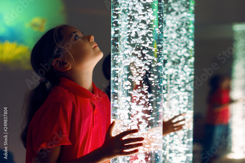 Child in therapy sensory stimulating room, snoezelen. Child interacting with colored lights bubble tube lamp during therapy session. photo