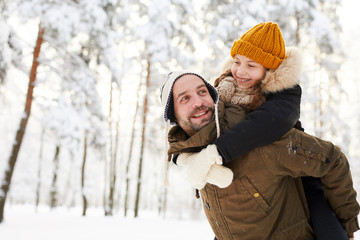 Portrait of happy little girl riding on fathers back while having fun in winter forest, copy space