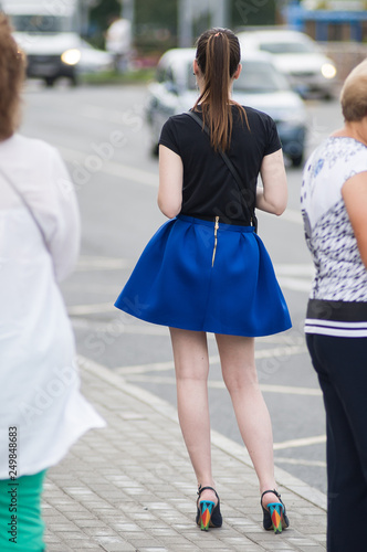 Beautiful young slender girl with long legs in blue short skirt stands at a public transport stop, the view from the back