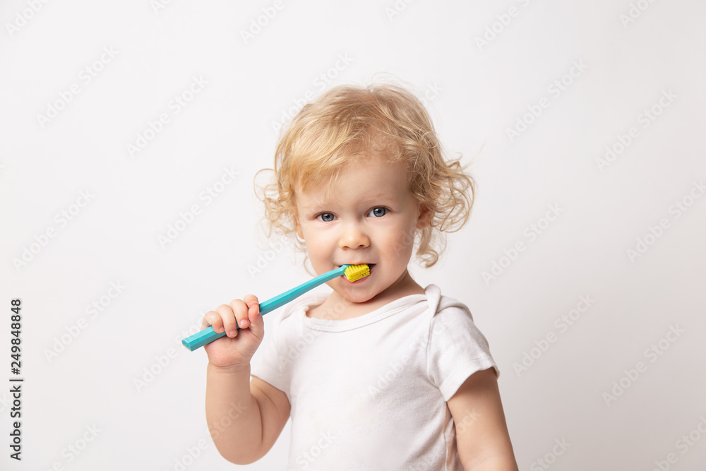 Caucasian little cute curly baby girl in white clothes body brushing her teeth with a colored blue brush on a white background. Caucasian little cute curly baby girl in white clothes body brushing her teeth with a colored blue brush on a white background.