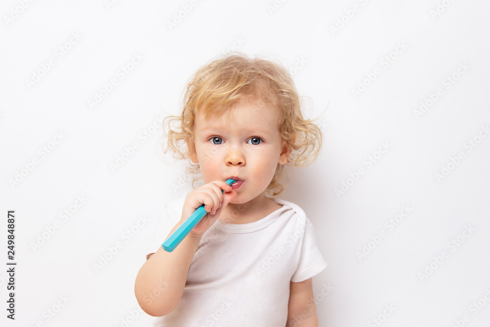 Caucasian little cute curly baby girl in white clothes body brushing her teeth with a colored blue brush on a white background. Caucasian little cute curly baby girl in white clothes body brushing her teeth with a colored blue brush on a white background.