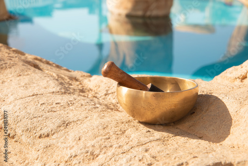 A singing Tibetan bowl stands on a sand-colored stone near a swimming pool on a sunny day. photo